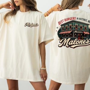 May include: Cream-coloured t-shirt with the text "Malone's" on the front and a graphic of a restaurant with the text "Best Burgers in Hastings, MA!" on the back. The restaurant graphic features a red and white striped awning.