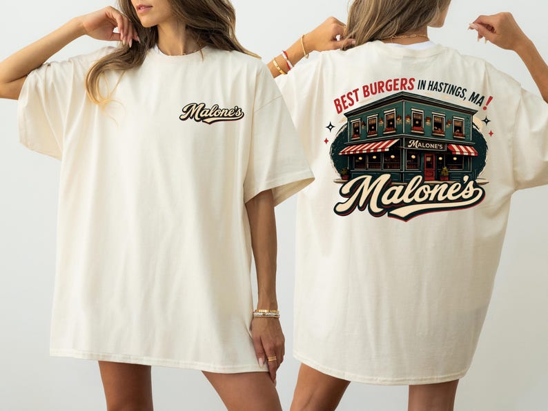 May include: Cream-coloured t-shirt with the text "Malone's" on the front and a graphic of a restaurant with the text "Best Burgers in Hastings, MA!" on the back. The restaurant graphic features a red and white striped awning.