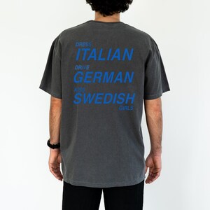 以下が含まれることがあります： 青い文字が入った濃い灰色のTシャツ。「DRESS ITALIAN DRIVE GERMAN KISS SWEDISH GIRLS」と書かれています。Tシャツは半袖でゆったりとしたフィット感です。黒いズボンを履いています。