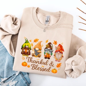Puede incluir: Sudadera beige con un gráfico colorido de cuatro gnomos sosteniendo calabazas, con hojas de otoño y el texto "Thankful & Blessed". Los gnomos visten varios sombreros y atuendos. Se ve un collar plateado.