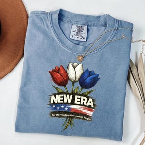 May include: A pale blue t-shirt featuring a floral design of red, white, and blue tulips. The text "NEW ERA" is above a banner with stars and stripes, and the text "For the Freedom of the Iranian People" is below. A gold necklace is draped across the shirt.
