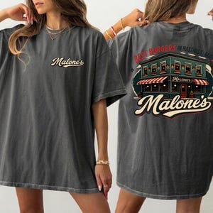 May include: Dark grey t-shirt with the Malone's logo on the front and a restaurant graphic on the back, with the text "Best Burgers in Hastings, MA!". The shirt has a vintage, worn appearance.
