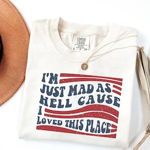 May include: A cream-colored short-sleeve t-shirt with a Comfort Colors brand label at the neck. The front features a graphic with red wavy stripes and blue text that reads: I'M JUST MAD AS HELL CAUSE I LOVED THIS PLACE. A gold necklace rests on the shirt.