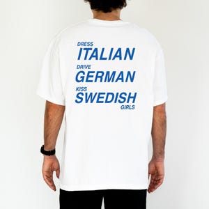 以下が含まれることがあります： 背中に青い文字が入った白いTシャツ。「DRESS ITALIAN DRIVE GERMAN KISS SWEDISH GIRLS」と書かれています。このシャツはカジュアルなスタイルです。