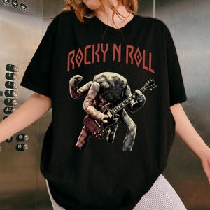 May include: Black t-shirt with the text "ROCKY N ROLL" in red. The shirt features a graphic of a creature playing a guitar. The creature is in shades of grey and brown.