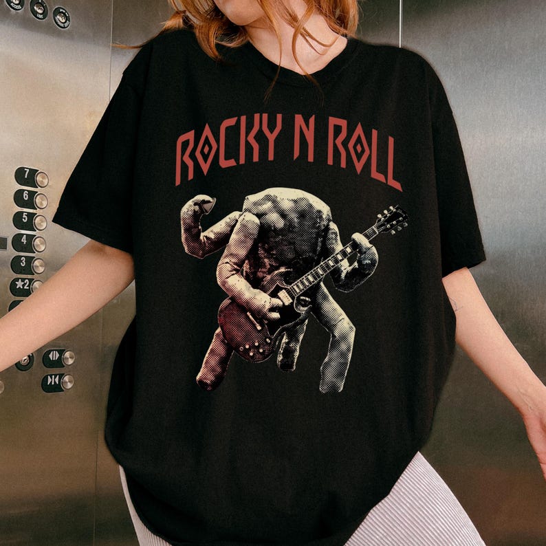 May include: Black t-shirt with the text "ROCKY N ROLL" in red. The shirt features a graphic of a creature playing a guitar. The creature is in shades of grey and brown.