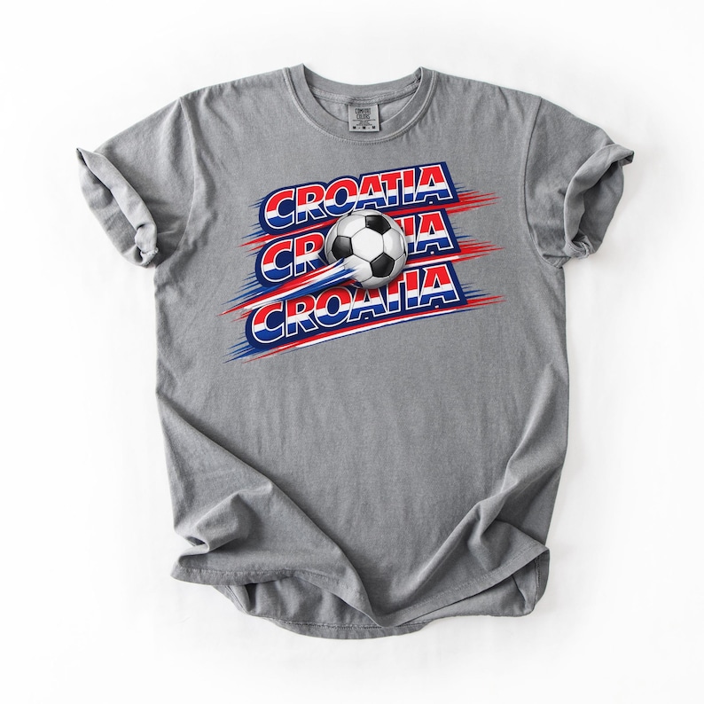 May include: A gray t-shirt featuring the word "Croatia" in red, white, and blue, with a soccer ball graphic. The design has a dynamic, sporty feel, with the word "Croatia" repeated three times.