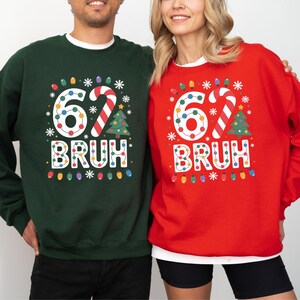 Op de afbeelding: Twee mensen dragen bijpassende sweatshirts. De ene is donkergroen, de andere rood. Beide hebben het nummer "62" met kerstverlichting, een zuurstok, een kerstboom en het woord "BRUH" in een feestelijk lettertype.