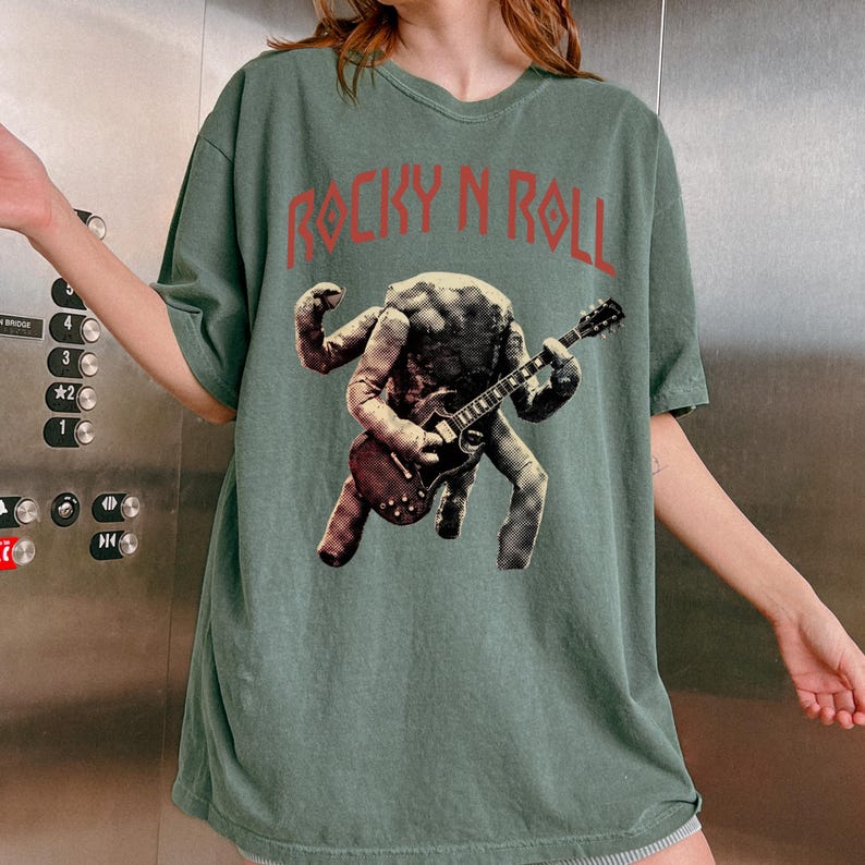 May include: A sage green t-shirt with the text "ROCKY N ROLL" in red. The shirt features a graphic of a figure playing a guitar. The figure has a textured appearance, and the guitar is brown.