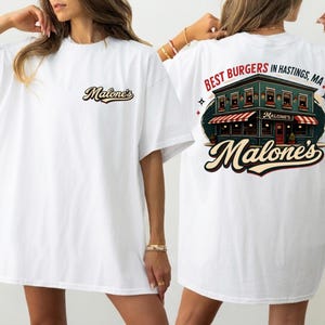 May include: White t-shirt with the text "Malone's" on the front and a graphic on the back. The back graphic features a building with a red and white striped awning, the words "BEST BURGERS IN HASTINGS, MA!" and the name "Malone's".