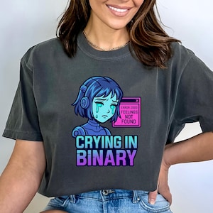May include: Dark grey t-shirt featuring a crying anime-style character with blue hair and headphones. The graphic includes the text "Crying in Binary" in a gradient font, and an error message that reads "Feelings Not Found."