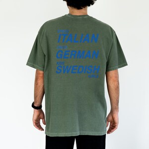 以下が含まれることがあります： オリーブグリーンのTシャツに青い文字。テキストは「Dress Italian, Drive German, Kiss Swedish Girls」と書かれています。このシャツはカジュアルスタイルで、半袖です。