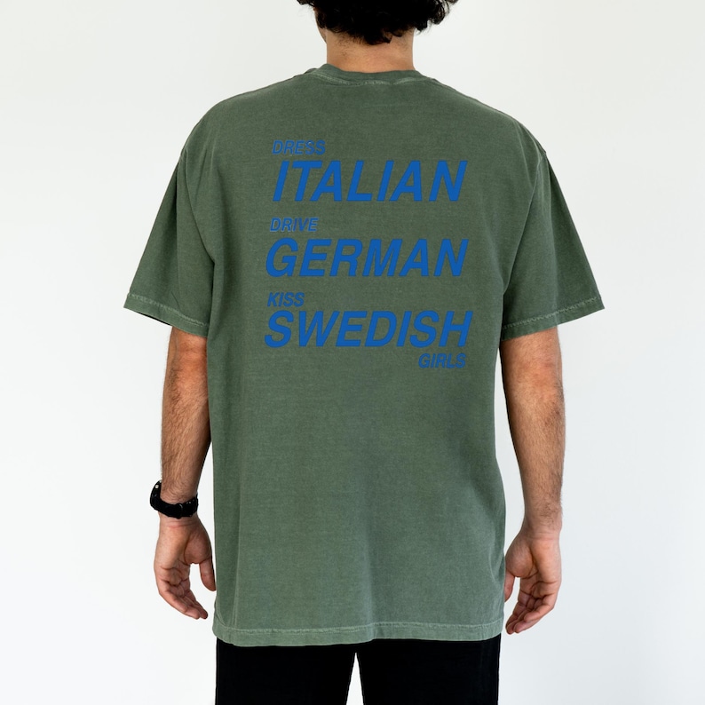 以下が含まれることがあります： オリーブグリーンのTシャツに青い文字。テキストは「Dress Italian, Drive German, Kiss Swedish Girls」と書かれています。このシャツはカジュアルスタイルで、半袖です。