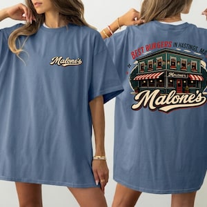 May include: A blue t-shirt with the Malone's logo on the front and a restaurant graphic on the back, with the text "Best Burgers in Hastings, MA!". The shirt has a loose fit.