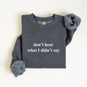 Puede incluir: Una sudadera gris oscuro con el texto blanco "don't hear what I didn't say". La sudadera de cuello redondo tiene mangas largas, con los puños enrollados. La tela parece suave y cómoda.