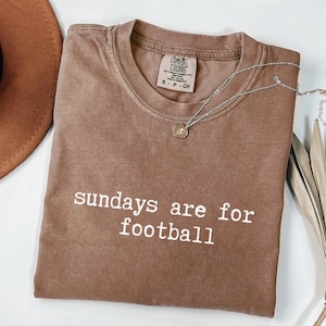 May include: A brown Comfort Colors t-shirt with the text "sundays are for football" in white. A silver necklace with a pendant is draped across the shirt. A brown hat is visible in the upper left corner.