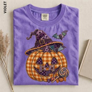 Puede incluir: Una camiseta morada con un gráfico de una calabaza de Halloween con sombrero de bruja, decorada con un patrón de cuadros y lunares. El diseño incluye dos pequeños murciélagos y una piruleta. La camiseta tiene una etiqueta Comfort Colors.