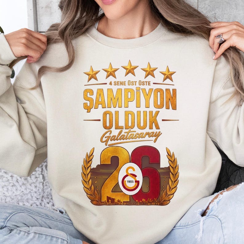 Puede incluir: Sudadera color crema con un dise&ntilde;o gr&aacute;fico. El dise&ntilde;o presenta estrellas doradas, el texto "4 SENE &Uuml;ST &Uuml;STE", "ŞAMPİYON OLDUK Galatasaray", y el n&uacute;mero "26" con un logotipo y el a&ntilde;o "1905".