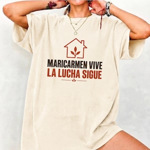 May include: A cream-coloured short-sleeve t-shirt featuring a brown graphic of a house outline with a plant inside. Below the graphic, the text reads MARICARMEN VIVE LA LUCHA SIGUE in dark brown and orange lettering with a small decorative plant icon.