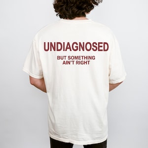 Puede incluir: Camiseta color crema con el texto "UNDIAGNOSED" en letras de bloque granates sobre la frase "BUT SOMETHING AIN'T RIGHT". La camiseta es de estilo casual, probablemente de algodón o tejido similar.
