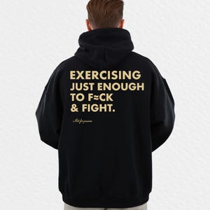 May include: Black hooded sweatshirt with the text "EXERCISING JUST ENOUGH TO F**K & FIGHT" in gold lettering. Below the text is the phrase "Not for pus**es" in a smaller font. The sweatshirt has a drawstring hood and a kangaroo pocket.