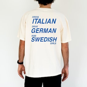 以下が含まれることがあります： 背中に青い文字がプリントされたクリーム色のTシャツ。「DRESS ITALIAN DRIVE GERMAN KISS SWEDISH GIRLS」と書かれています。このシャツはカジュアルなスタイルで、おそらく綿または同様の生地で作られています。