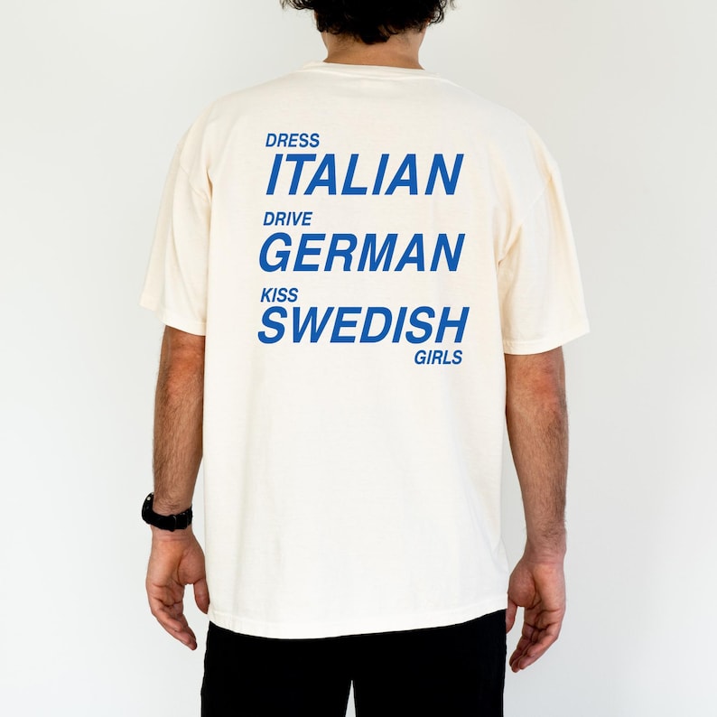 以下が含まれることがあります： 背中に青い文字がプリントされたクリーム色のTシャツ。「DRESS ITALIAN DRIVE GERMAN KISS SWEDISH GIRLS」と書かれています。このシャツはカジュアルなスタイルで、おそらく綿または同様の生地で作られています。