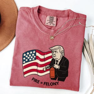 May include: A dusty rose Comfort Colors t-shirt featuring a graphic of a man holding a fire extinguisher in front of an American flag. The text "FIRE = FELONY" is below the graphic. A gold necklace is draped across the shirt.