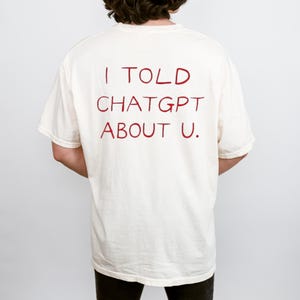 Puede incluir: Camiseta color crema con el texto "I TOLD CHATGPT ABOUT U." en rojo, con letras manuscritas. La camiseta es de estilo casual, de manga corta. La imagen muestra la parte trasera de la camiseta.