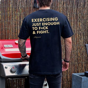 May include: Black t-shirt with gold text that reads "EXERCISING JUST ENOUGH TO F*CK & FIGHT." The shirt also has the text "Not for pu**ies" below the main text. The shirt is being worn by a person standing near a red grill.