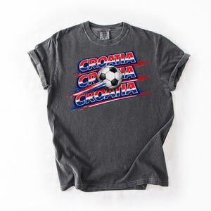 May include: Dark gray t-shirt featuring the word "CROATIA" in red, white, and blue, with a soccer ball graphic. The design is repeated three times with a motion effect. The shirt is made of a soft material.