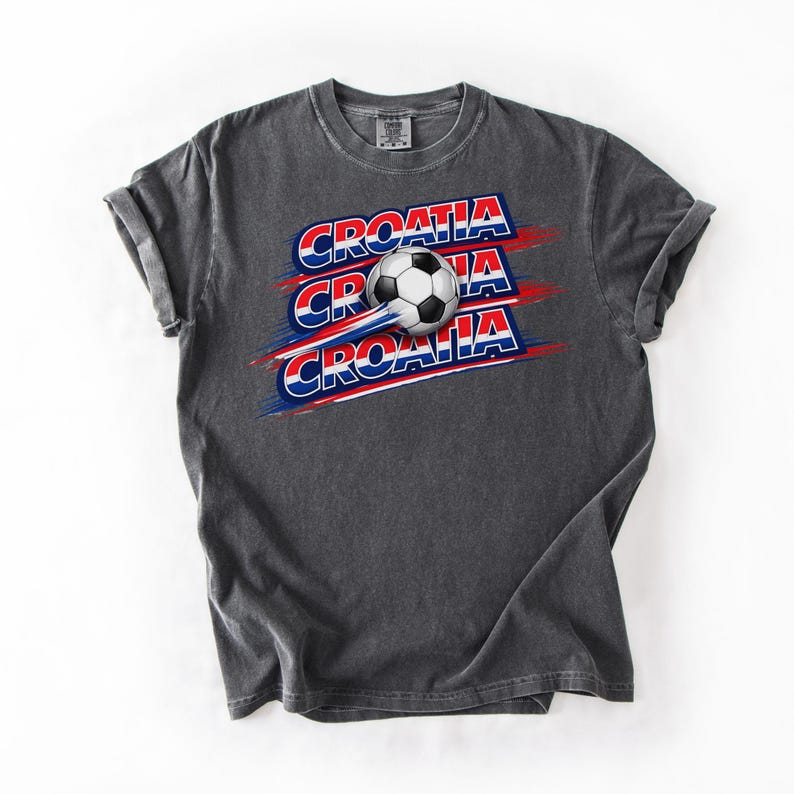 May include: Dark gray t-shirt featuring the word "CROATIA" in red, white, and blue, with a soccer ball graphic. The design is repeated three times with a motion effect. The shirt is made of a soft material.