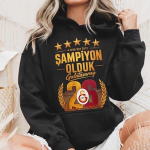 Puede incluir: Sudadera con capucha negra con texto dorado y detalles de estrellas. El texto dice "4 SENE &Uuml;ST &Uuml;STE ŞAMPİYON OLDUK Galatasaray 26 1905". El dise&ntilde;o incluye un emblema dorado y rojo con el n&uacute;mero 26.