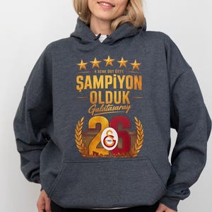 Puede incluir: Sudadera con capucha gris oscuro con un gr&aacute;fico dorado y rojo. El gr&aacute;fico incluye texto y el n&uacute;mero 26, con el logotipo del Galatasaray. El texto dice "4 SENE &Uuml;ST &Uuml;STE ŞAMPİYON OLDUK Galatasaray".