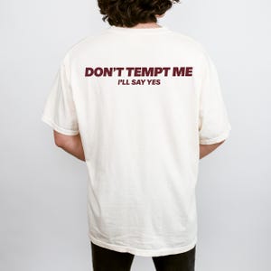 May include: Cream-colored t-shirt with the text "DON'T TEMPT ME I'LL SAY YES" printed in a dark burgundy color across the upper back. The shirt has short sleeves and a relaxed fit.