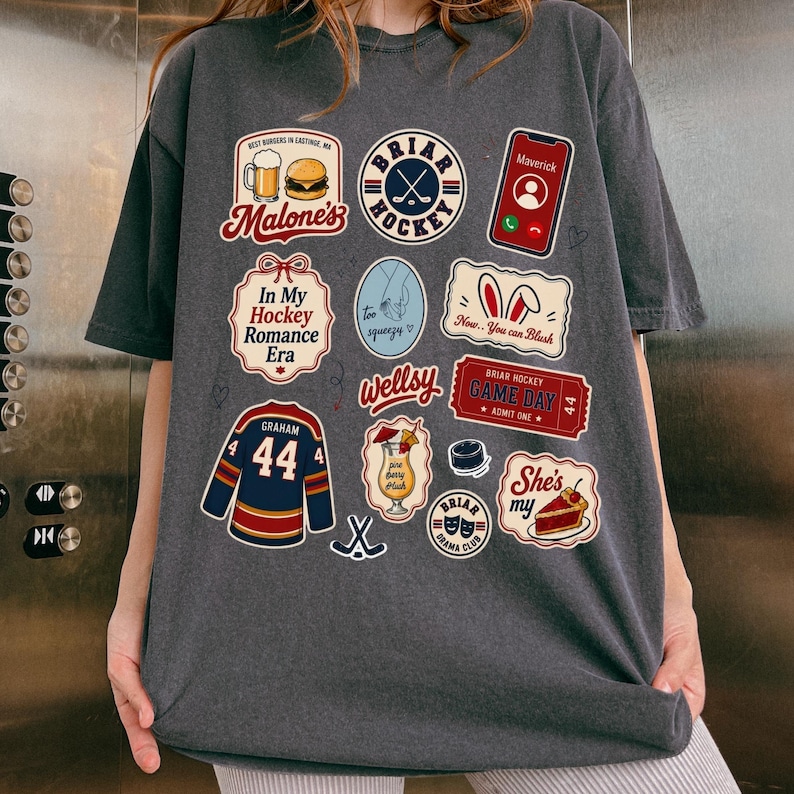 Op de afbeelding: Donkergrijs T-shirt met diverse kleurrijke grafische patches. Patches bevatten tekst als "Briar Hockey", "Malone's" en "In My Hockey Romance Era". Andere patches tonen een hockeytrui, een cocktail en een telefoon.