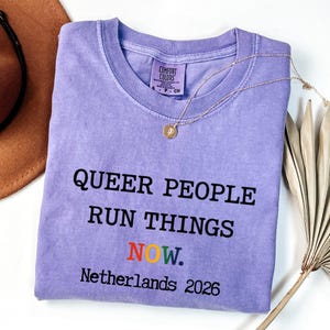 以下が含まれることがあります： 「QUEER PEOPLE RUN THINGS NOW. Netherlands 2026」のテキストが黒色で、虹色の「NOW」がプリントされた、ラベンダー色のComfort Colors Tシャツ。 金色のネックレスと茶色の帽子も写っています。