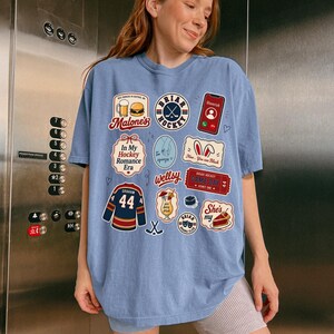 Op de afbeelding: Lichtblauw T-shirt met diverse grafische patches. Patches zijn onder meer een hockeylogo, een telefoon met een oproep, een hockeytrui met nummer 44 en tekst met de tekst "Malone's", "In My Hockey Romance Era" en "She's my".