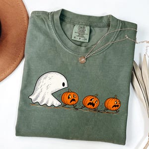 Puede incluir: Una camiseta de manga corta color verde oliva con un gráfico de un fantasma blanco persiguiendo a tres calabazas naranjas con expresiones faciales angustiadas. La camiseta tiene cuello redondo y una etiqueta Comfort Colors en el interior.