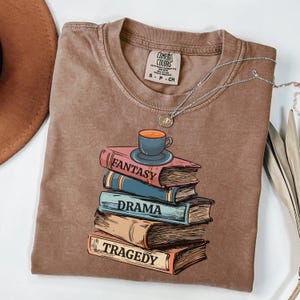 May include: A brown t-shirt with a graphic of a stack of books with the words "Fantasy", "Drama", and "Tragedy" on the spines. A small cup of coffee sits on top of the books.
