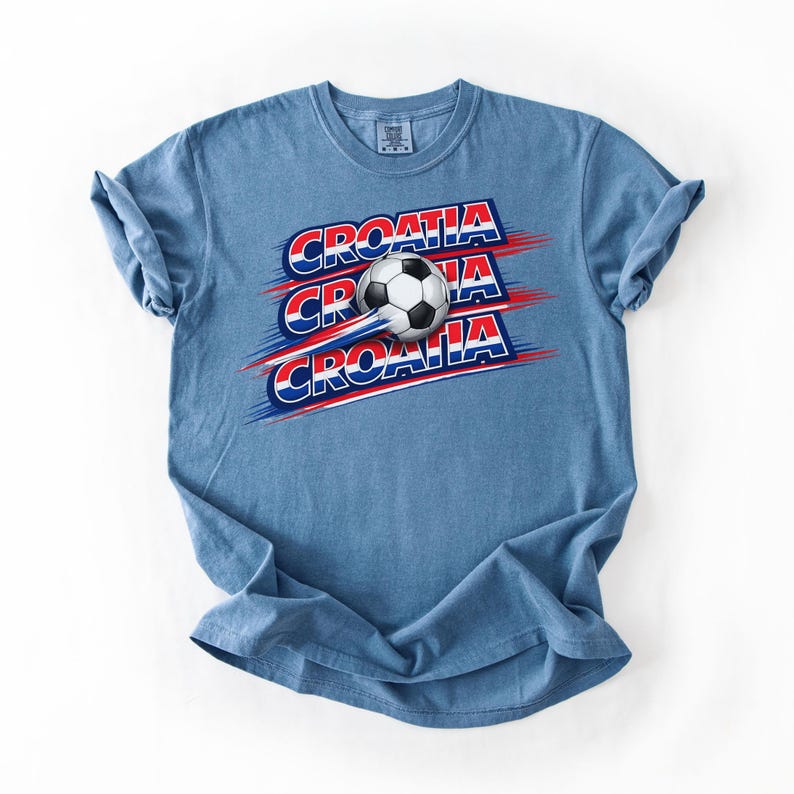 May include: A blue t-shirt featuring the word "CROATIA" in red and white, with a soccer ball graphic. The design includes red and blue streaks, suggesting motion. The shirt is a casual wear item.
