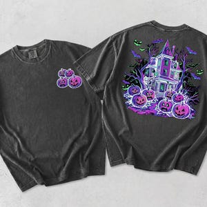 Puede incluir: Una camiseta gris carbón de manga corta con un pequeño gráfico de cuatro calabazas moradas en el pecho izquierdo y un gráfico grande y detallado en la espalda de una casa encantada rodeada de calabazas moradas, árboles desnudos y murciélagos.