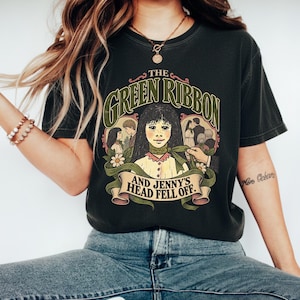 May include: A charcoal gray short-sleeve t-shirt featuring a graphic illustration of a person with a green ribbon around their neck. The text reads THE GREEN RIBBON AND JENNY'S HEAD FELL OFF, surrounded by floral accents and smaller character vignettes.