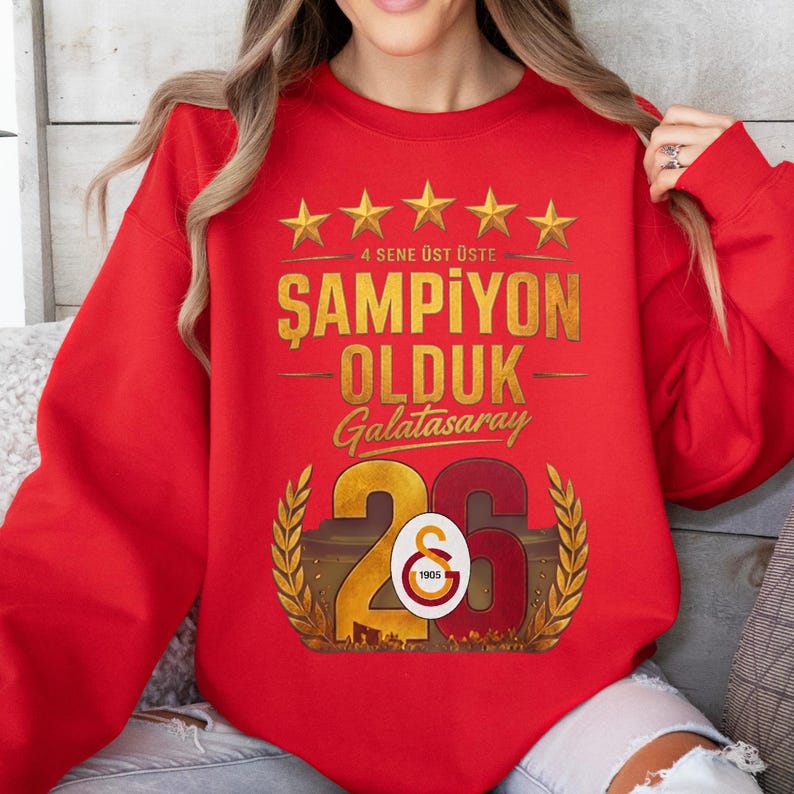Puede incluir: Una sudadera roja con texto y gr&aacute;ficos dorados. El texto dice "4 SENE &Uuml;ST &Uuml;STE ŞAMPİYON OLDUK Galatasaray 26". Encima del texto hay cinco estrellas doradas. El n&uacute;mero 26 es en oro y rojo.