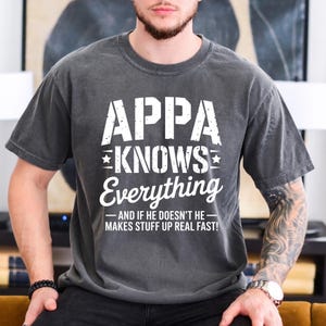May include: Dark gray t-shirt with the text "APPA KNOWS Everything - AND IF HE DOESN'T HE - MAKES STUFF UP REAL FAST!" in white. The shirt is a casual wear item.