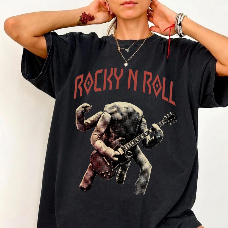 May include: Black t-shirt with the text "ROCKY N ROLL" in red. The shirt features a graphic of a figure playing a guitar. The figure has multiple arms and a textured appearance, giving it a vintage rock and roll vibe.