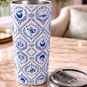 May include: A stainless steel tumbler with a white and blue pattern featuring apples, books, and other academic symbols. The tumbler has a silver rim and a clear lid. The design is reminiscent of Delftware pottery.