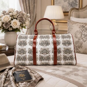 May include: A white travel bag featuring a grey rose pattern and brown leather handles. The bag is rectangular and rests on a bed with a floral scarf and a passport.