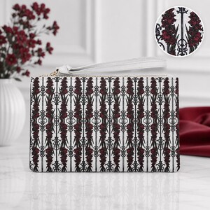 May include: A rectangular wristlet clutch featuring a white background with a repeating vertical pattern of dark red floral stalks and black wrought iron-style scrollwork. The bag includes a white wrist strap and a gold-toned zipper closure.