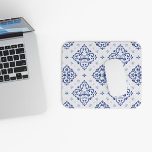 May include: A white mouse mat featuring a blue and white floral pattern, with a white computer mouse resting on it. The mouse mat has the words "TEACH", "INSPIRE", and "GUIDE". A laptop is in the background.